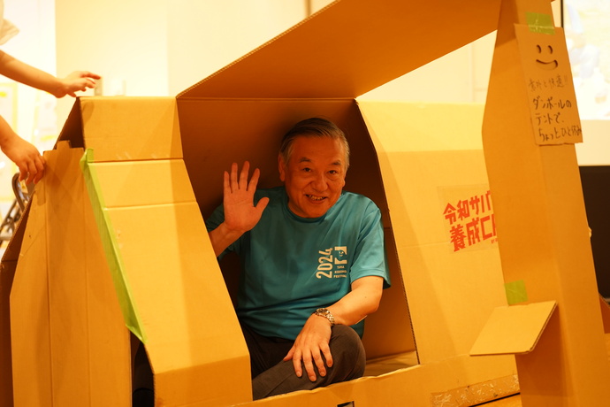 ダンボールテントに入って手を振る多摩市長